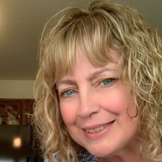 A person with curly blonde hair smiling at the camera, indoors. The background includes part of a room with a refrigerator and a piece of art on the wall.