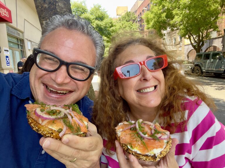 Two people smiling and holding bagels topped with smoked salmon, cream cheese, onions, and herbs. They are wearing glasses and posing outdoors on a sunny day with trees and buildings in the background.