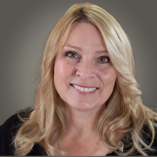A person with long blond hair smiling at the camera, wearing a black top, set against a gray background.