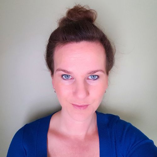 A person with light skin and blue eyes is looking directly into the camera. They have their brown hair tied up in a high bun and are wearing a blue top. The background is a plain, light-colored wall.