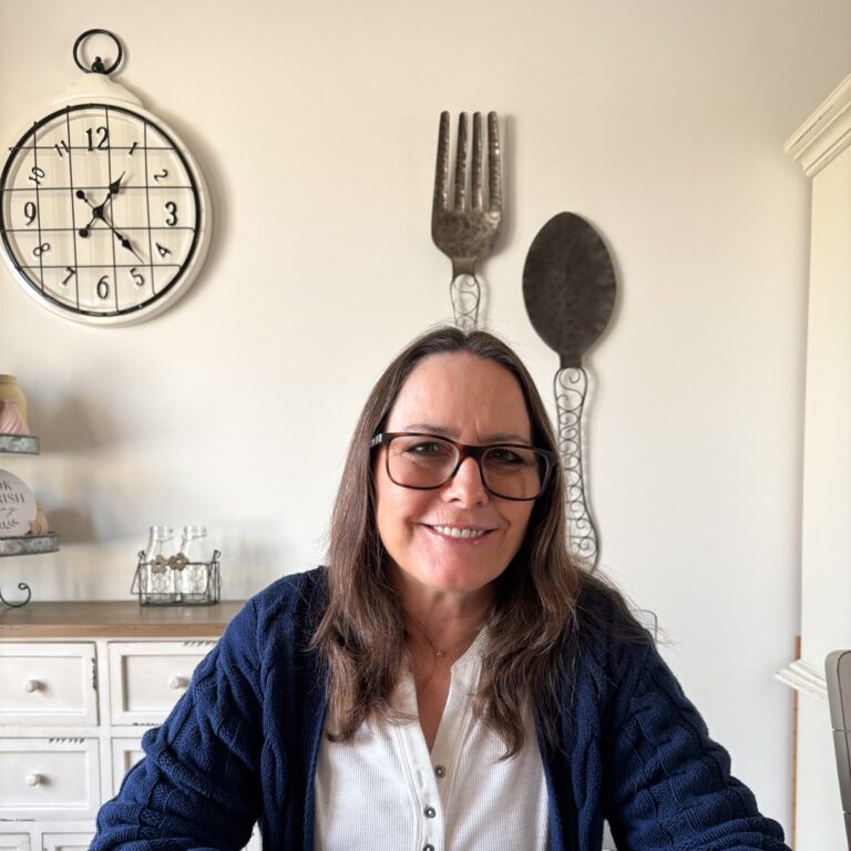 A person with long brown hair and glasses smiles while sitting indoors. They wear a navy cardigan over a white blouse. Behind them are large decorative wall utensils and a clock, with a cabinet adorned with small items to the left.