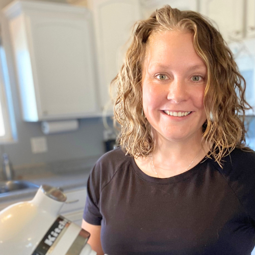 A person with wavy hair smiles while standing in a kitchen. They are wearing a black shirt and holding a KitchenAid mixer. The kitchen has white cabinets and light walls, with a window visible in the background.