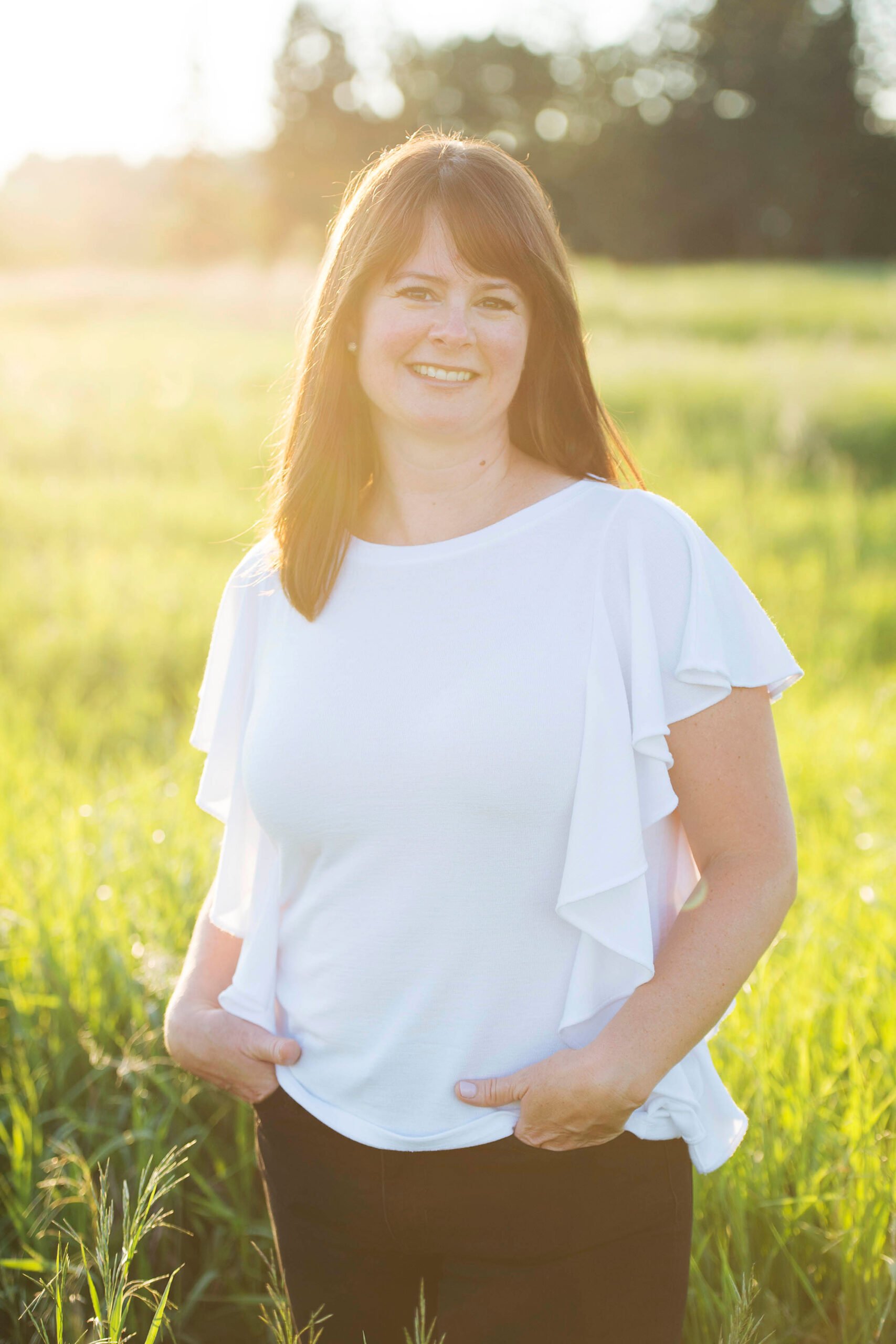 A woman with long brown hair stands in a sunlit field, wearing a white blouse and black pants. She smiles warmly, with her hands in her pockets. Green grass and blurred trees create a serene background.