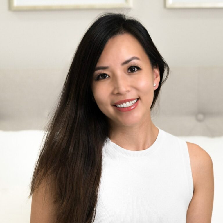 Person with long dark hair and a white sleeveless top smiling in a softly lit setting, with a blurred light-colored background.