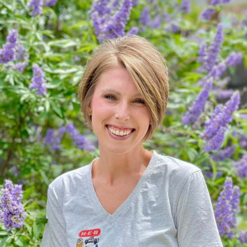A person with short hair smiling outside, wearing a light gray H-E-B shirt. They are standing in front of a lush background with vibrant purple flowers and green foliage.