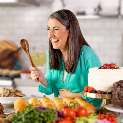 A woman in a turquoise blouse laughs joyfully while holding a wooden spoon. She's surrounded by a variety of foods, including bread rolls, a decorated cake, and colorful vegetables, in a bright kitchen setting.