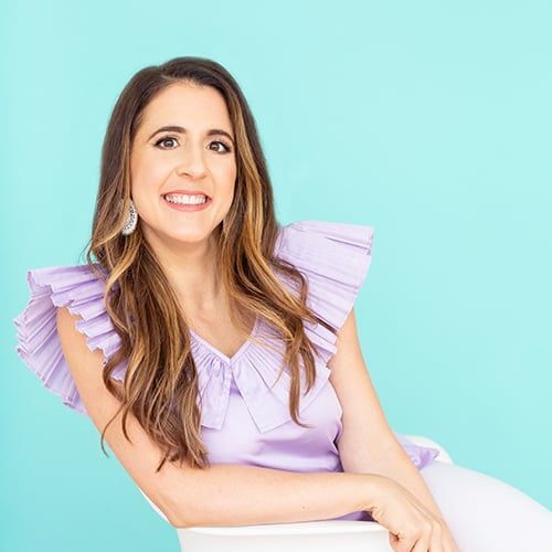 A woman with long brown hair is smiling and leaning on a white chair. She is wearing a lavender top with ruffled sleeves and silver earrings. The background is a solid light teal color.