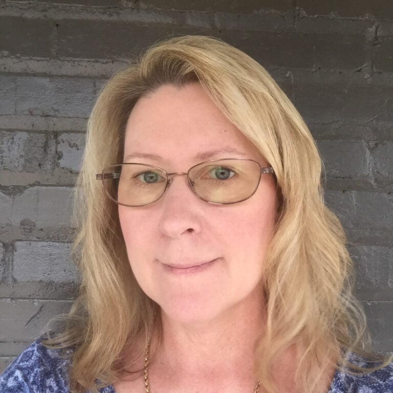 A woman with shoulder-length blonde hair and glasses stands in front of a gray brick wall. She is wearing a blue and white patterned top and a gold necklace.