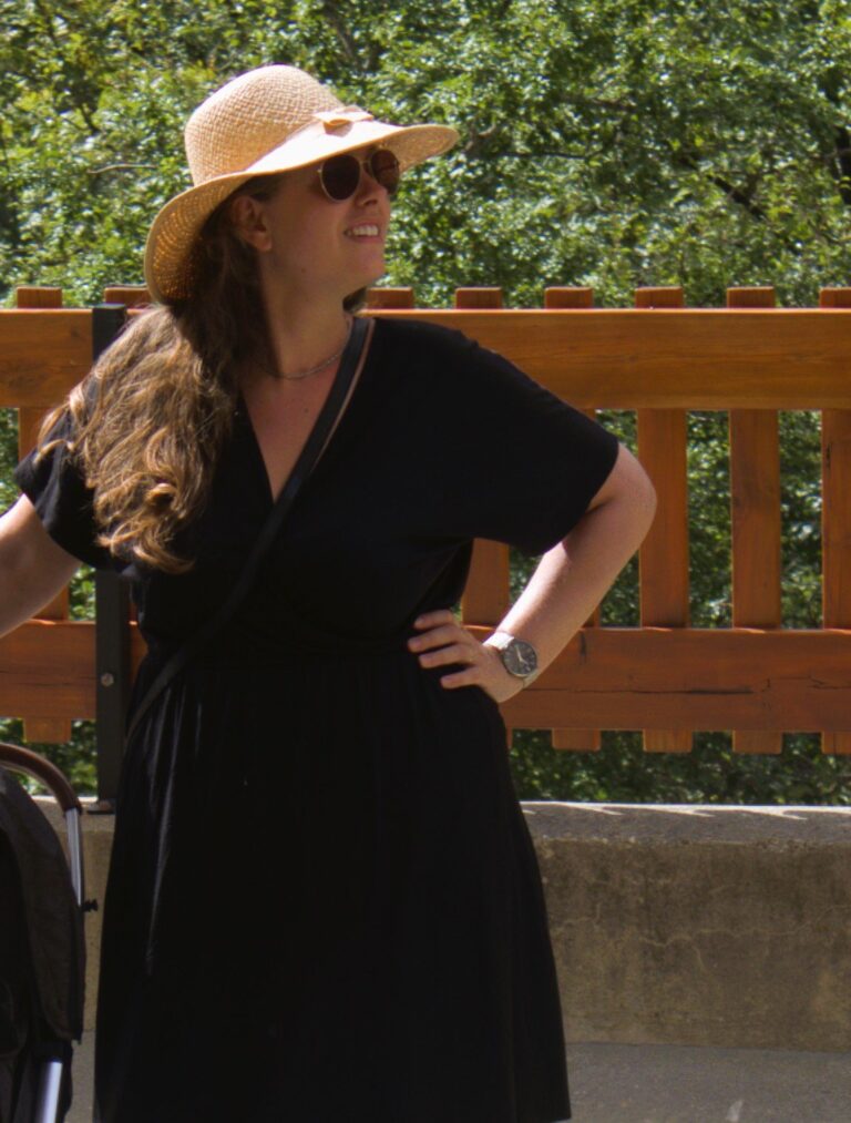 A woman in a black dress and straw hat stands outdoors, looking to the side. She wears sunglasses and a watch, with her hand on her hip. A stroller is partially visible beside her. A wooden fence and greenery are in the background.