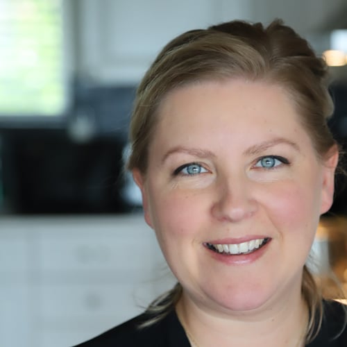A person with light hair smiling warmly at the camera. They are standing in a kitchen with blurred cabinets in the background, creating a soft and inviting atmosphere.