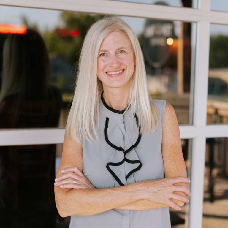 A smiling woman with long, straight blonde hair stands with her arms crossed in front of a glass door. She is wearing a sleeveless grey blouse with a black trim and ruffle detail. The background depicts a blurred outdoor scene.