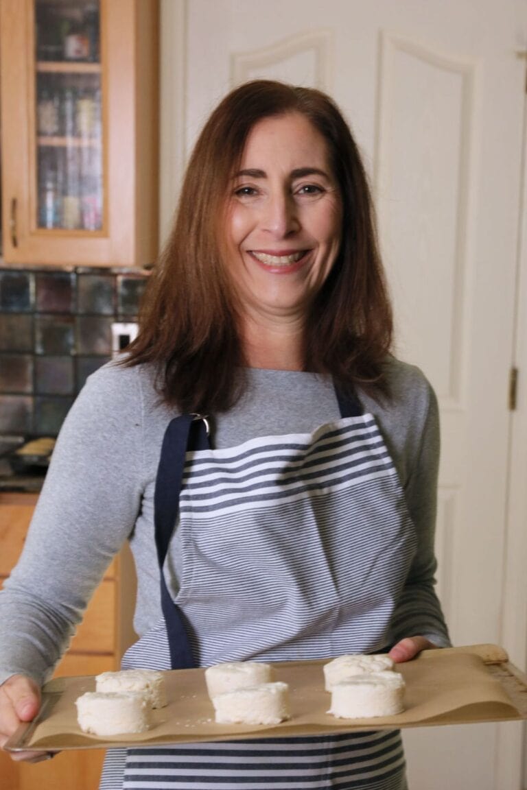 A smiling woman with long brown hair wearing a gray shirt and a striped apron holds a baking tray with uncooked dough pieces. She is standing in a kitchen with wooden cabinets and tiled backsplash.