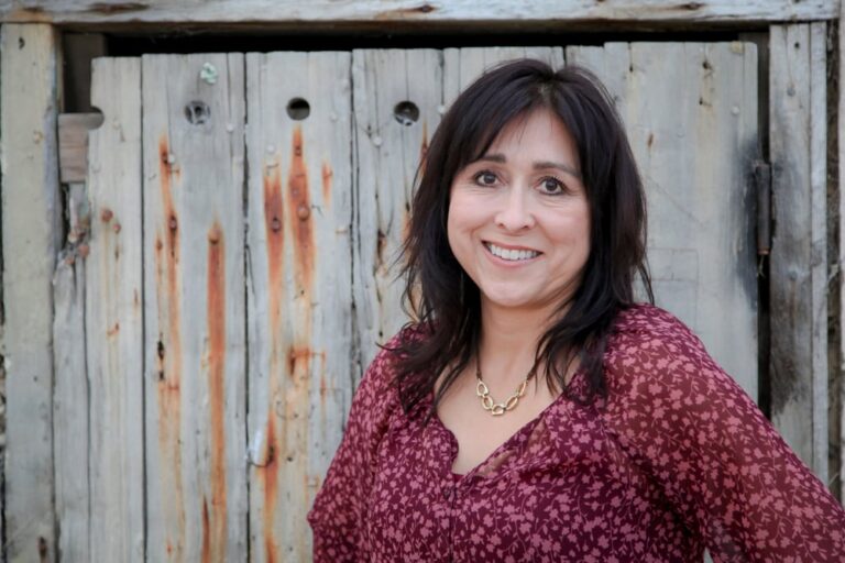Woman with dark hair wearing a maroon patterned blouse stands smiling in front of a weathered wooden fence.