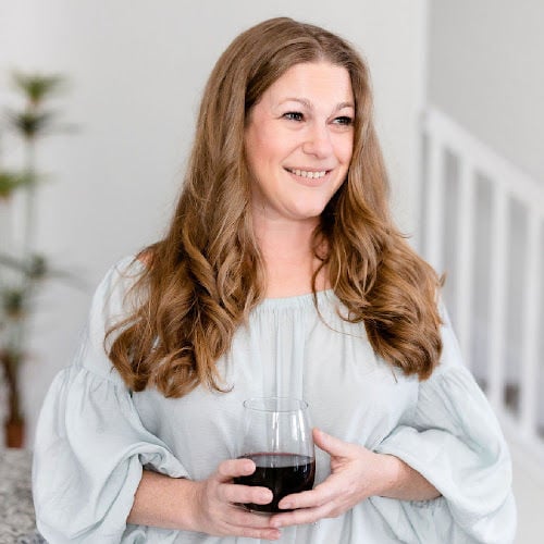 A woman with long hair smiles while holding a glass of red wine. She is wearing a light-colored, off-the-shoulder top. The background features a blurred staircase and plants.