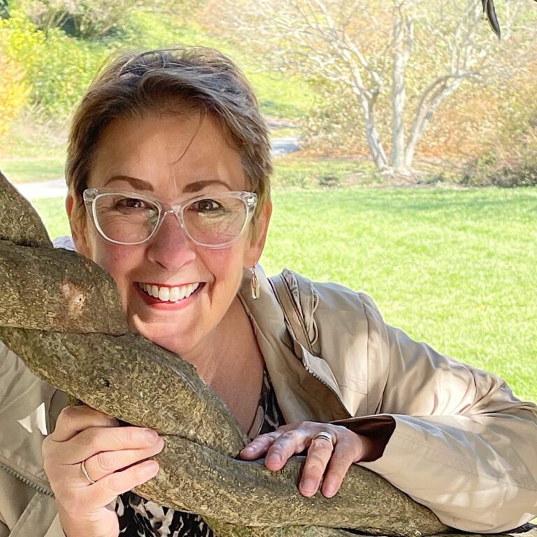 A smiling woman with short hair and glasses stands outdoors, holding onto a tree branch. She's wearing a beige jacket and patterned shirt. The background features a grassy area and light-colored trees.