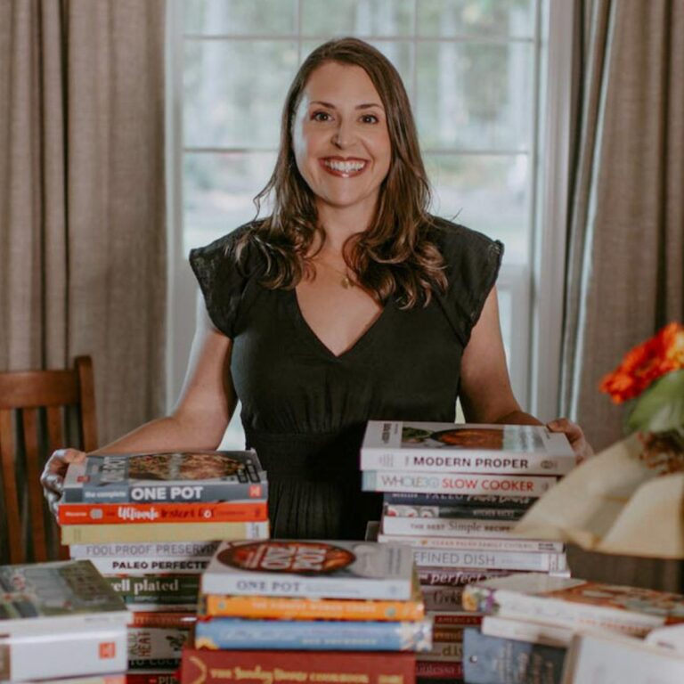 A woman in a black dress stands smiling behind a table piled with cookbooks. She is in a room with large windows and a curtain in the background.