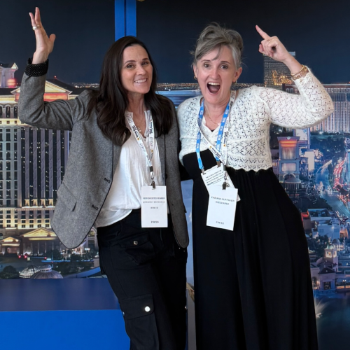 Two women smiling and posing with their arms raised in front of a backdrop depicting a cityscape. Both wear conference badges. One wears a gray jacket and black pants; the other wears a white and black outfit.