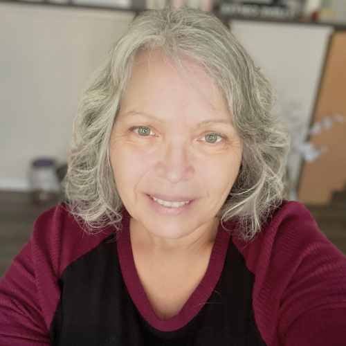 A woman with shoulder-length gray hair smiles at the camera. She is wearing a burgundy and black top. The background is softly blurred, featuring indoor elements.