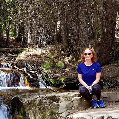 A woman in a blue shirt and sunglasses sits on a rock next to a flowing creek in a wooded area. Tall trees surround her, and the sunlight filters through the branches, creating a peaceful natural setting.