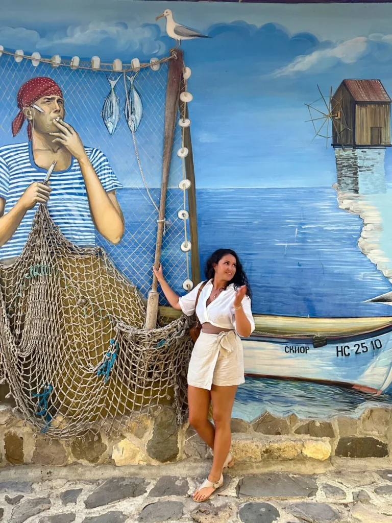 The Modern Nonna in front of a painting of a ship yard wearing a white romper.