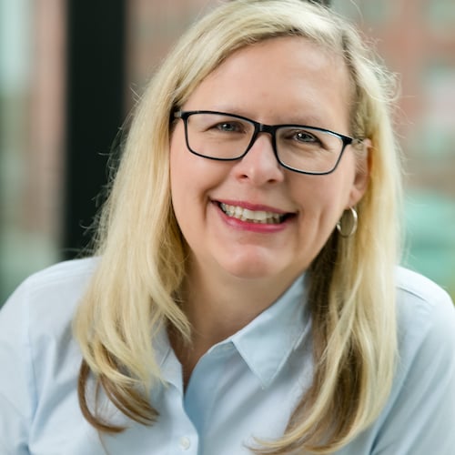 A smiling woman with long blonde hair wearing glasses. She is dressed in a light blue shirt and sitting indoors with a blurred window background.
