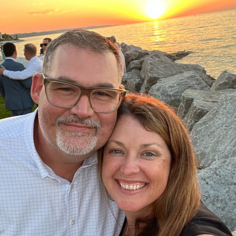 A smiling man and woman taking a selfie near large rocks by the water. The sun sets on the horizon, casting an orange glow on the ocean. Several people are in the background.