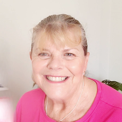 A smiling person with light hair is wearing a pink top. They are sitting indoors against a plain white wall, and there is a small plant in the background.