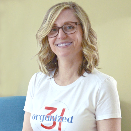 A woman with shoulder-length wavy blonde hair and glasses smiles at the camera. She is wearing a white T-shirt with the word "organized" and the number "31" printed on it in blue and red. She stands in front of a neutral background.