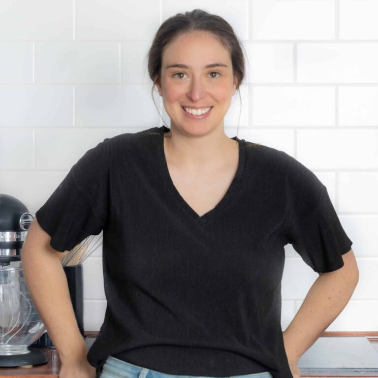 A person with dark hair tied back smiles at the camera in a kitchen. They are wearing a black V-neck shirt and jeans, with a white tiled wall and a countertop mixer in the background.