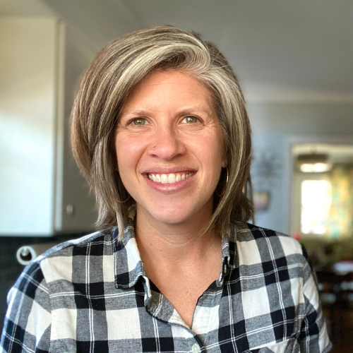 Woman with gray and white hair, smiling, wearing a black and white plaid shirt, in a well-lit room with blurred background.
