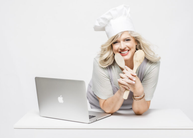 A smiling woman wearing a chef's hat holds two wooden spoons while leaning on a white surface with a laptop in front of her. She has blonde hair and is dressed in light-colored clothing. The background is white and clean.