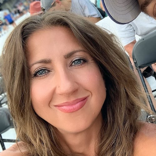 Smiling person with long brown hair, wearing light makeup, sits outdoors in a crowd with empty chairs in the background.