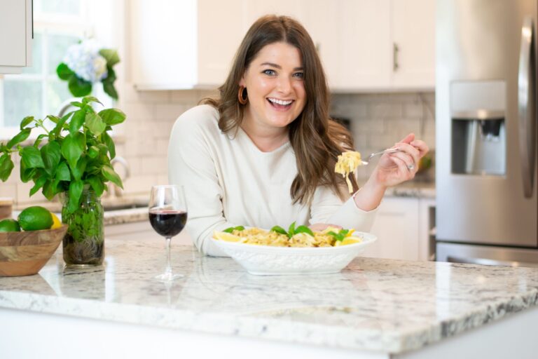 A woman smiles while sitting at a kitchen counter with a plate of pasta and a glass of red wine. A basil plant and a bowl of limes are beside her. She holds a forkful of pasta, enjoying the meal in a bright kitchen.