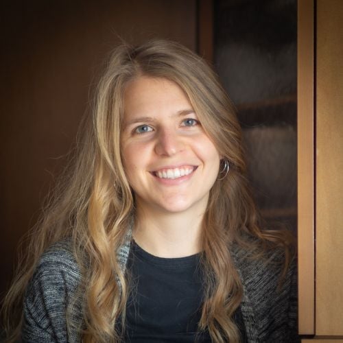Smiling person with long blond hair wearing a black shirt and gray cardigan, standing in front of a wooden cabinet.