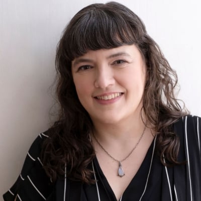 A person with long, wavy hair and bangs smiles at the camera. They are wearing a black and white striped top and a necklace with a teardrop-shaped pendant. The background is a plain, light-colored wall.