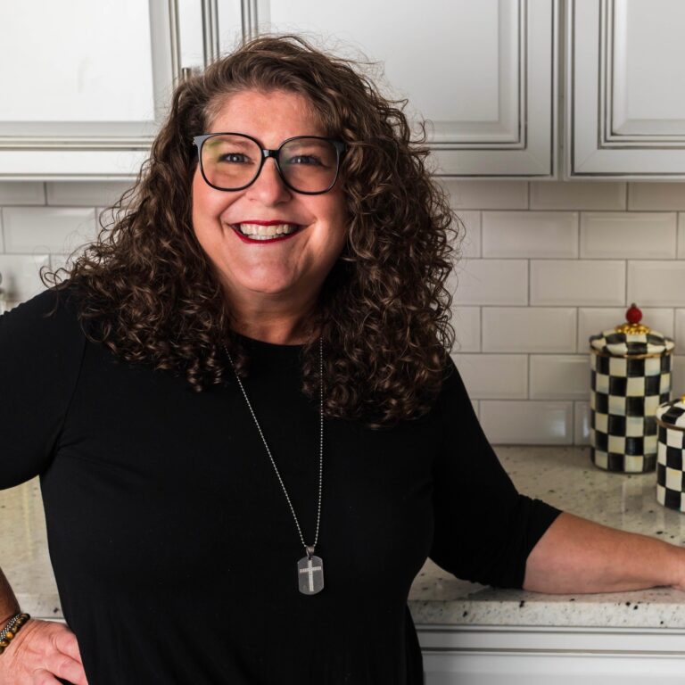 A woman with curly hair and glasses is smiling in a kitchen. She is wearing a black top and a necklace with a cross pendant. The kitchen has white cabinets and decorative items with checkerboard patterns on the counter.