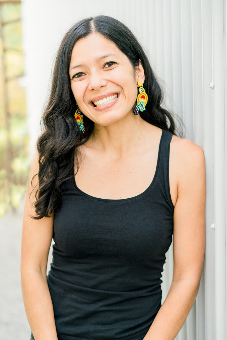 A woman with long dark hair smiles brightly while standing next to a corrugated metal wall. She is wearing a black tank top and colorful parrot-shaped earrings. Background is slightly out of focus, suggesting an outdoor setting.