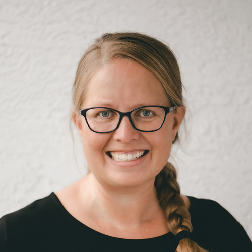 A smiling person with long, light brown braided hair and black-rimmed glasses, wearing a black top, stands against a light gray textured background.