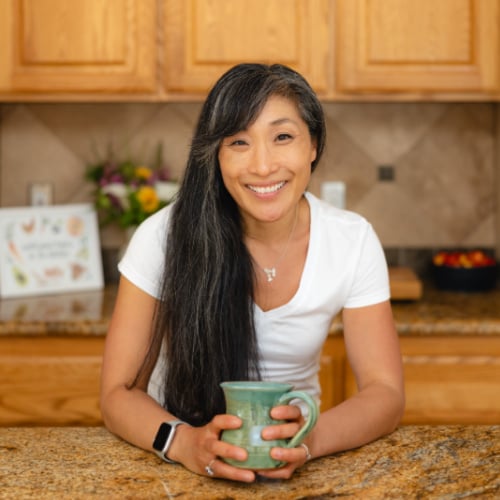 A person with long dark hair, wearing a white shirt and an Apple Watch, smiles while holding a green mug in a kitchen with wooden cabinets and a granite countertop. Flowers and a bowl of fruit are in the background.