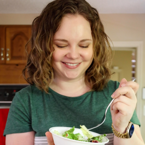 A smiling person with curly hair is holding a white bowl of salad and a fork. They are standing in a kitchen, wearing a green shirt, with an oven and wooden cabinets in the background.