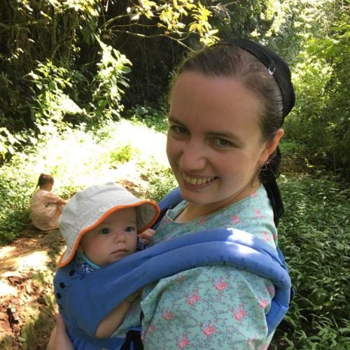 A woman smiles at the camera while carrying a baby in a blue carrier. The baby wears a white sun hat. They stand on a lush, green forest path with another child in the background.