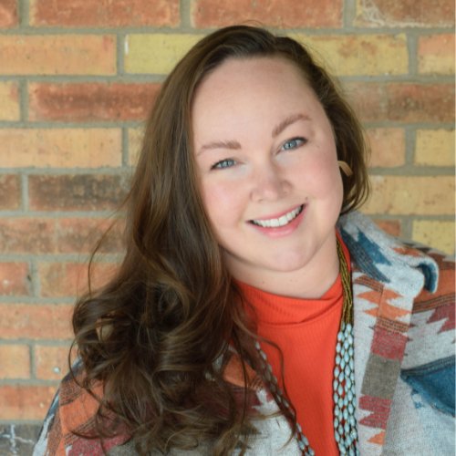 A woman with long brown hair and blue eyes smiles at the camera. She is wearing an orange top and a patterned shawl, standing in front of a brick wall.