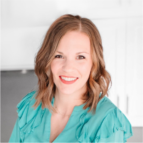 A person with shoulder-length wavy hair smiles at the camera. They are wearing a teal blouse and a delicate necklace. The background is softly blurred, featuring light-colored cabinets.