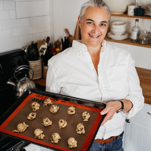 Person smiling and holding a baking tray filled with unbaked cookie dough balls in a kitchen. Wearing a white shirt, they stand near a stove with a kettle and shelves with dishes behind them.