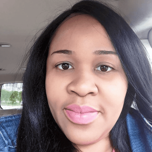 A woman with long black hair and light pink lipstick is inside a car. She is wearing a blue denim jacket and looking directly at the camera with a neutral expression. The car's interior is slightly visible in the background.