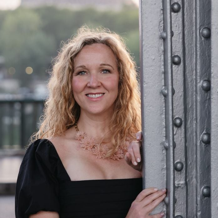 A woman with curly blonde hair smiles while standing outdoors. She's wearing a black dress with a floral necklace and is leaning against a metallic beam. Soft focus greenery is visible in the background.