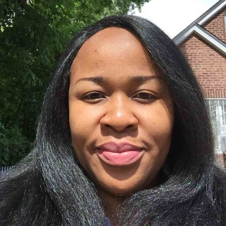 A person with long black hair smiling outdoors, with trees and a brick building in the background.