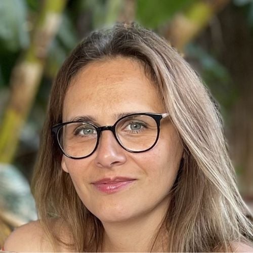 A person with long, light brown hair and glasses smiles at the camera. They are outdoors, with a blurred green background suggesting vegetation.