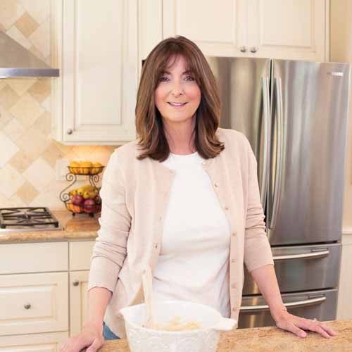 A woman with shoulder-length brown hair is standing in a kitchen, smiling. She is wearing a beige cardigan over a white shirt. There is a bowl with a wooden spoon on the counter in front of her. The kitchen has cream-colored cabinets and a stainless steel refrigerator.