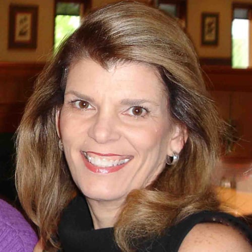 A woman with long brown hair and hoop earrings smiles warmly. She is wearing a black top and is in an indoor setting, with soft lighting and framed pictures in the background.